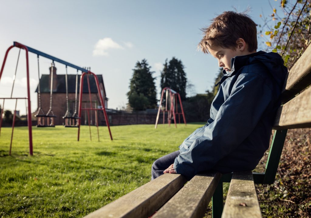 Lonely child sitting on play park playground bench Upset boy who has been bullied for bowel bladder leaks.